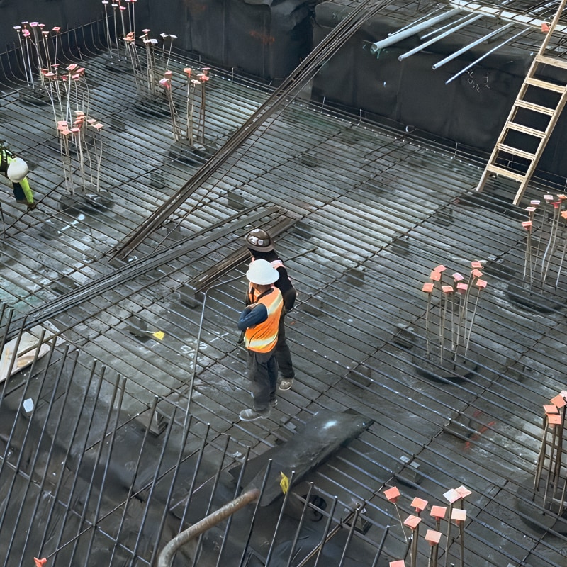 High-angle view of two construction workers wearing safety vests and hard hats, standing on a dense grid of steel rebar reinforcement mesh at a construction site.