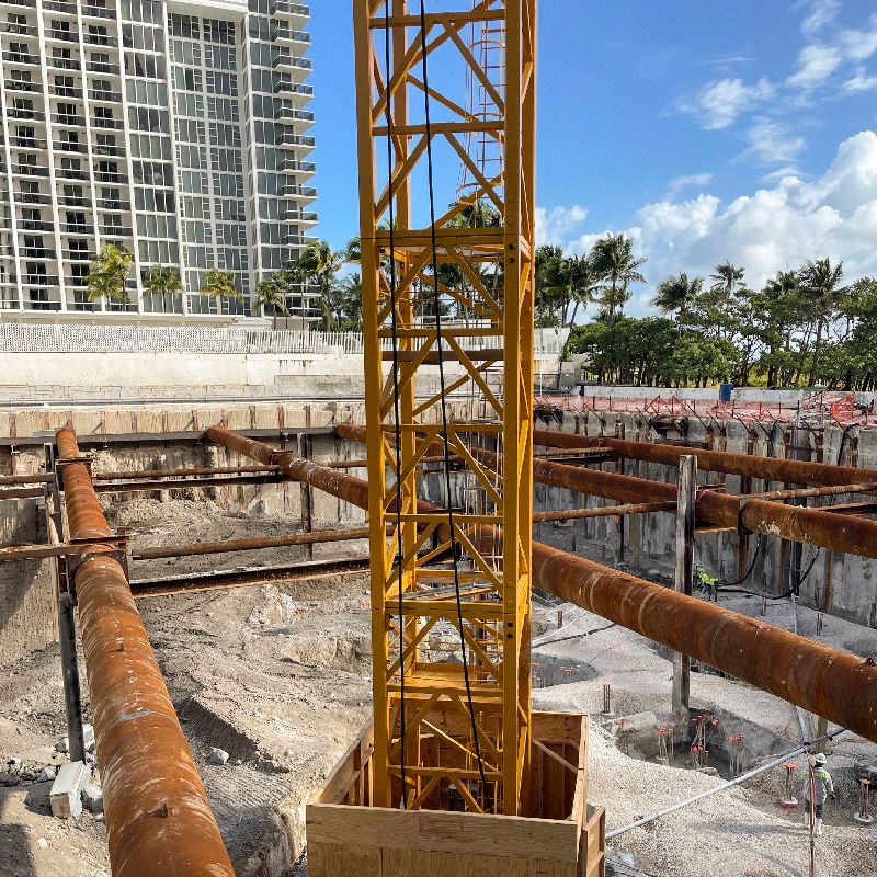 A yellow tower crane mast stands in the foreground of a deep foundation construction excavation site, featuring large rusty steel pipe struts and a high-rise building and palm trees in the background under a blue sky.