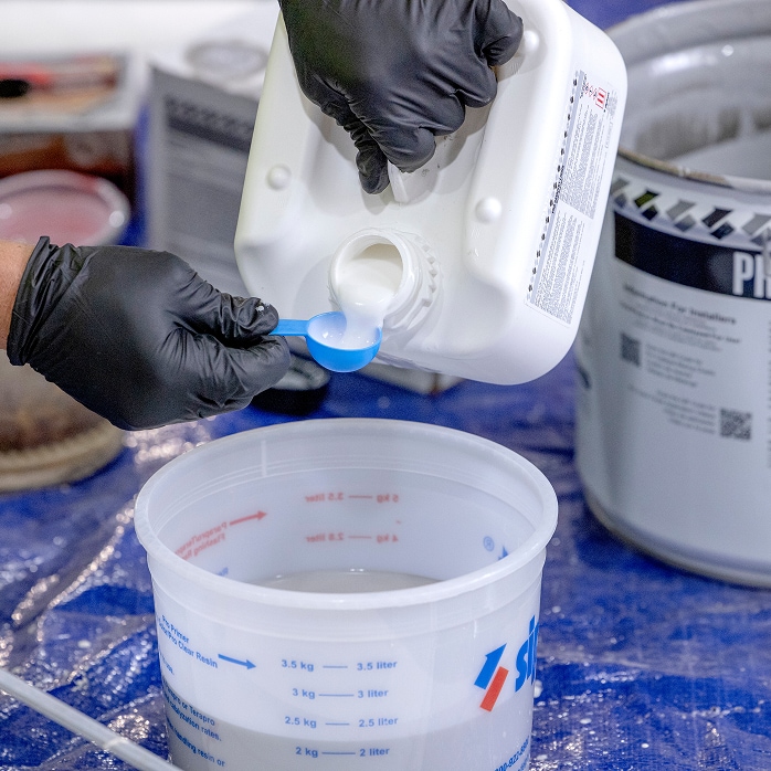 Gloved hands mixing liquid catalyst into a container, illustrating material estimating guides and calculators for liquid products.