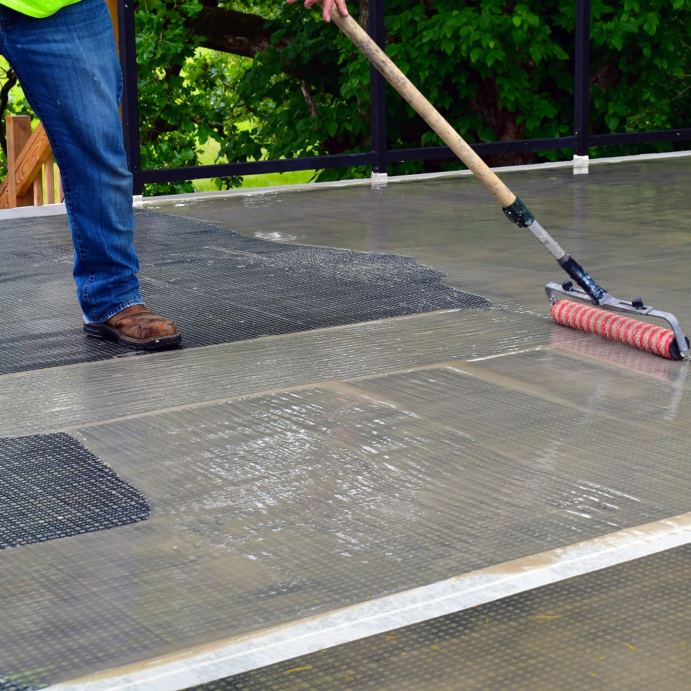 Worker applying PMMA liquid waterproofing resin with a roller over a reinforced plaza membrane.