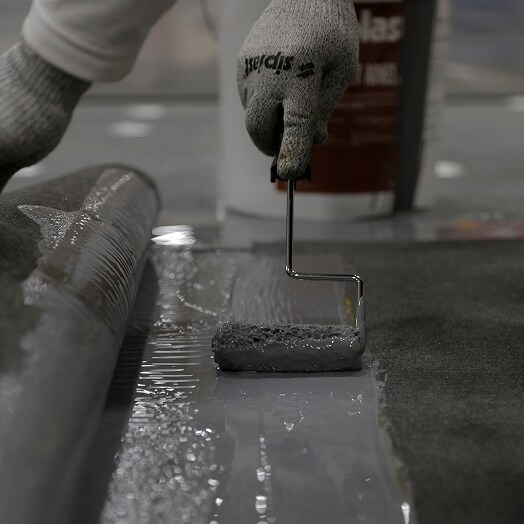 A worker applying adhesive with a roller to a Paradiene 30 modified bitumen roofing top ply membrane.