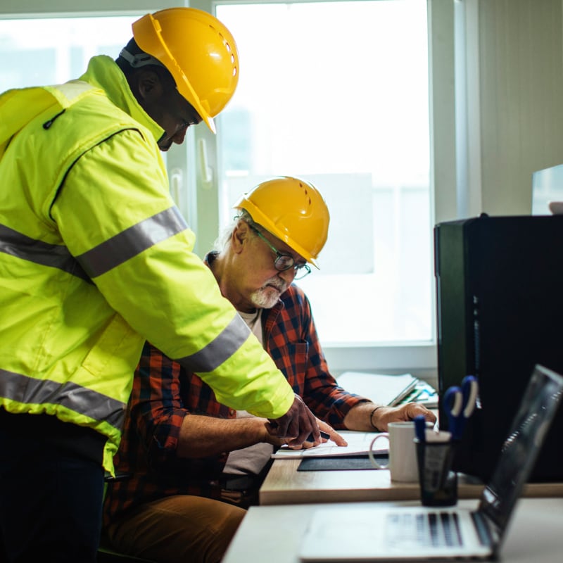Two construction workers wearing hard hats and safety vests are collaborating over a laptop and blueprints on a desk indoors, suggesting design guidance and technical support