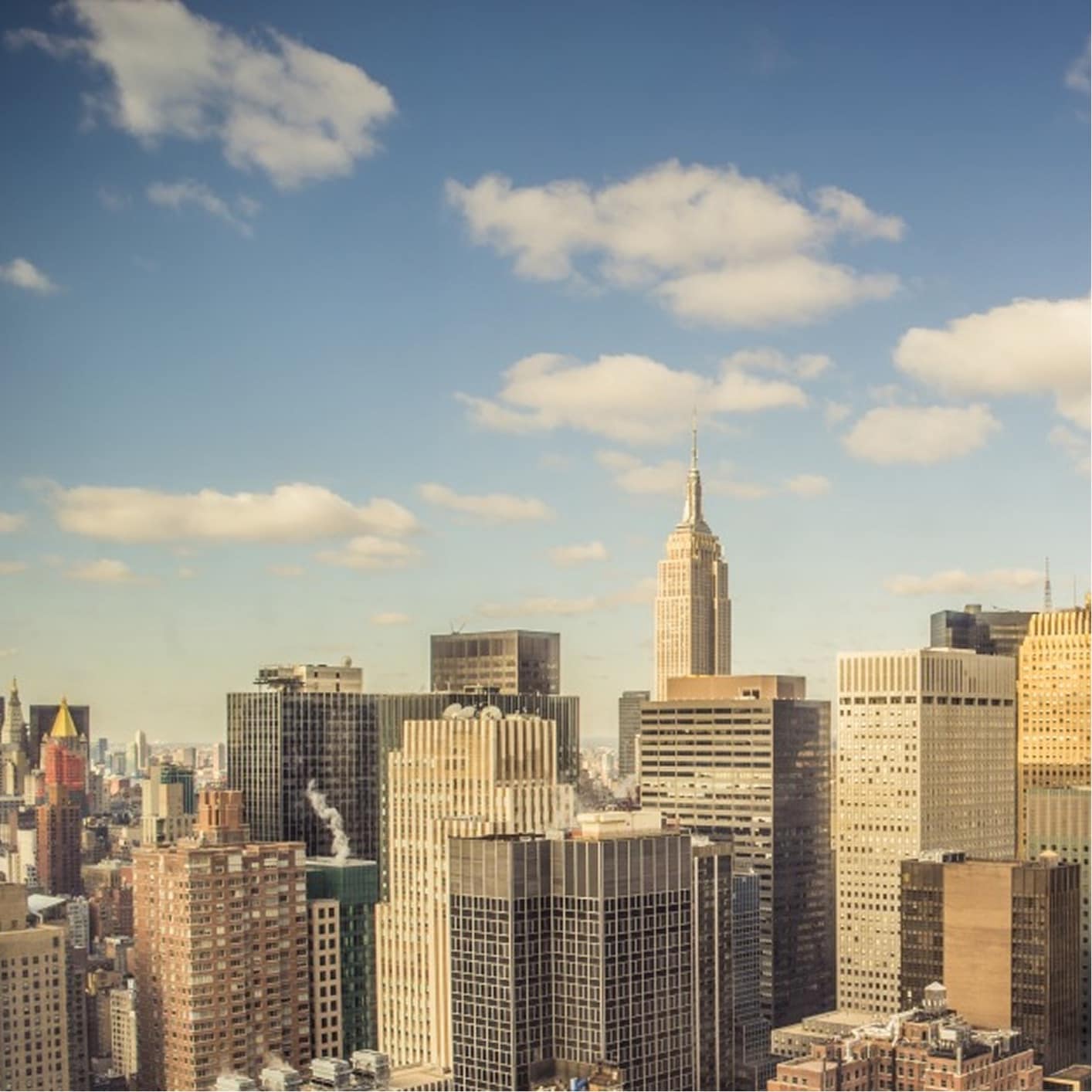 Panoramic view of the New York City skyline on a clear day, featuring iconic skyscrapers like the Chrysler Building and Empire State Building, under a partly cloudy sky.