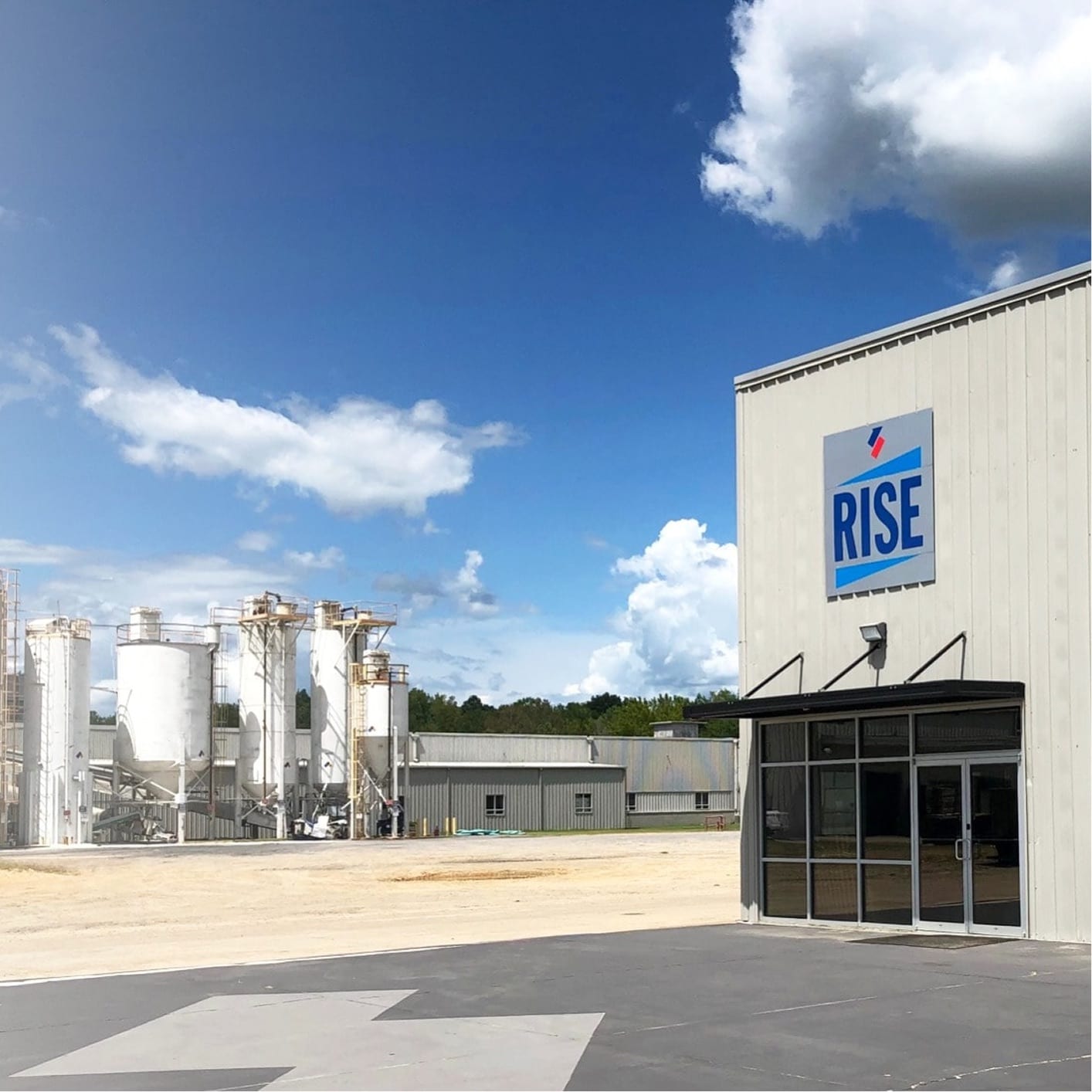 Exterior view of a large industrial facility with a RISE company logo on the building, featuring silos and manufacturing infrastructure under a blue sky with clouds.