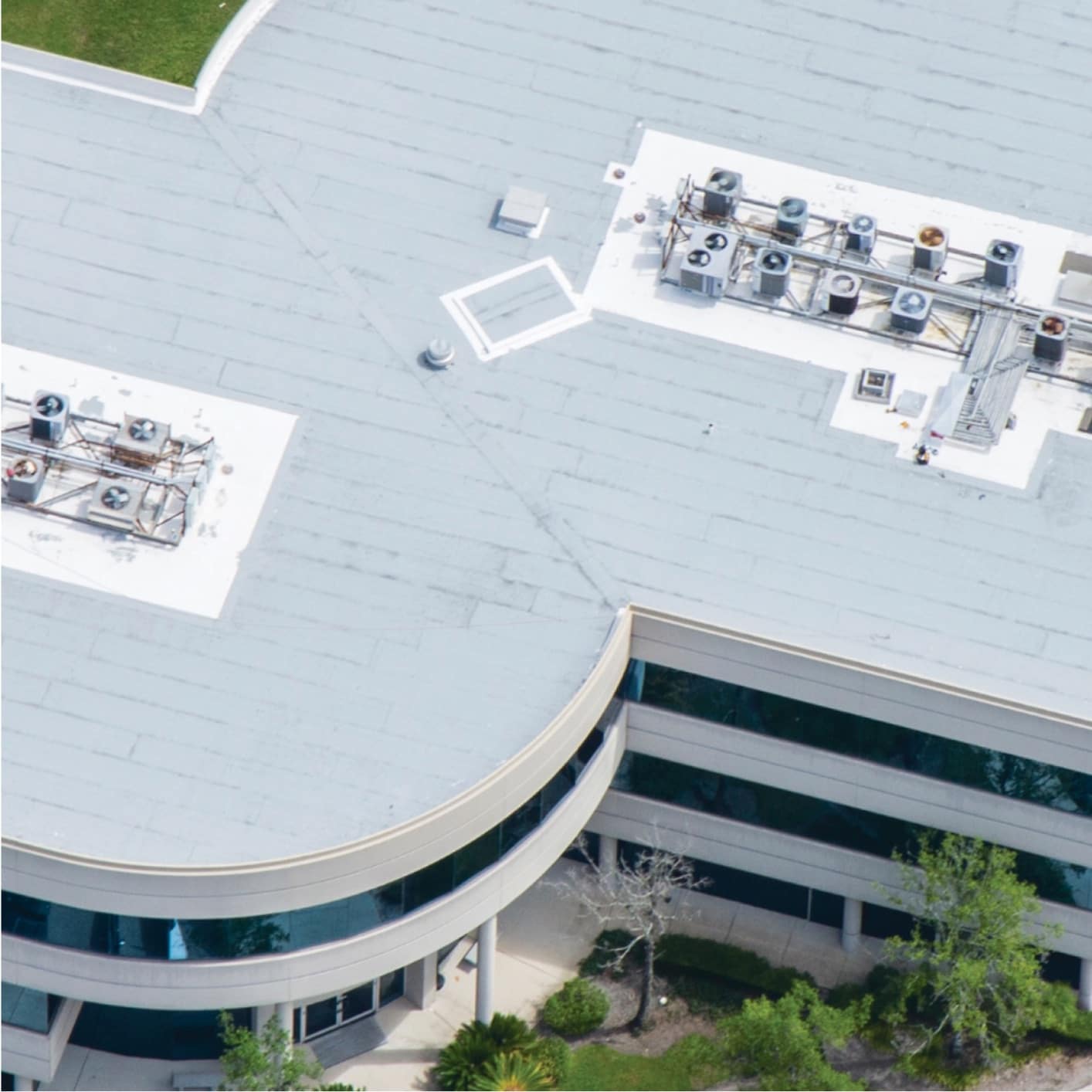 Aerial view of a contemporary office building with a light-colored flat roof, featuring multiple Siplast PMMA and surrounded by green trees and a parking lot.