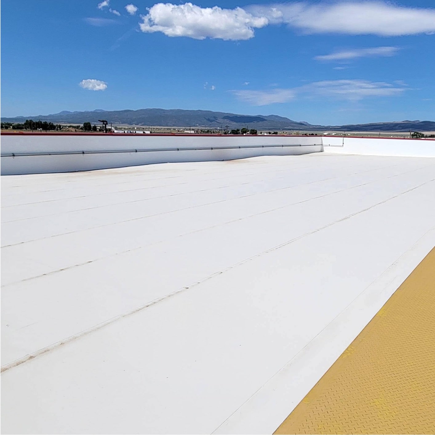 Expansive white TPO commercial roofing system installed on a large flat roof, with a yellow walkway path and distant mountains under a clear blue sky.