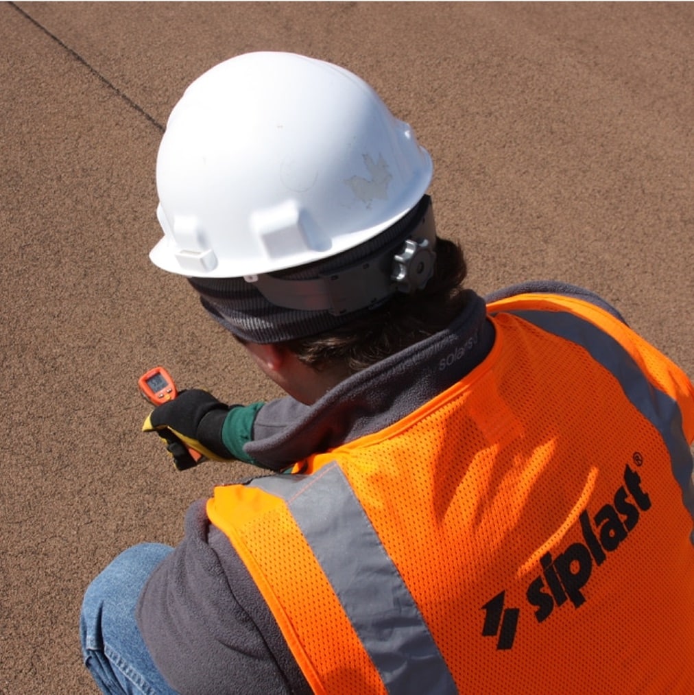A roofing professional in an orange Siplast safety vest and white hard hat uses a handheld tool to inspect the temperature of a brown Siplast commercial roof, demonstrating Siplast's commitment to robust roofing and waterproofing systems.