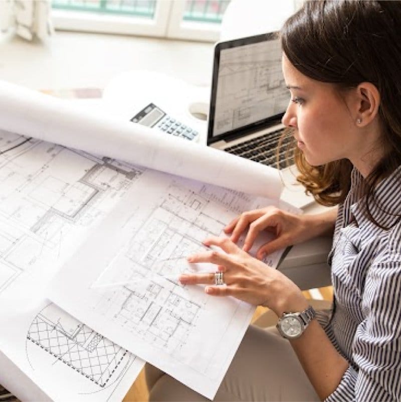 A female design professional reviewing detailed architectural blueprints and using a laptop, illustrating Siplast resources for design and specification.