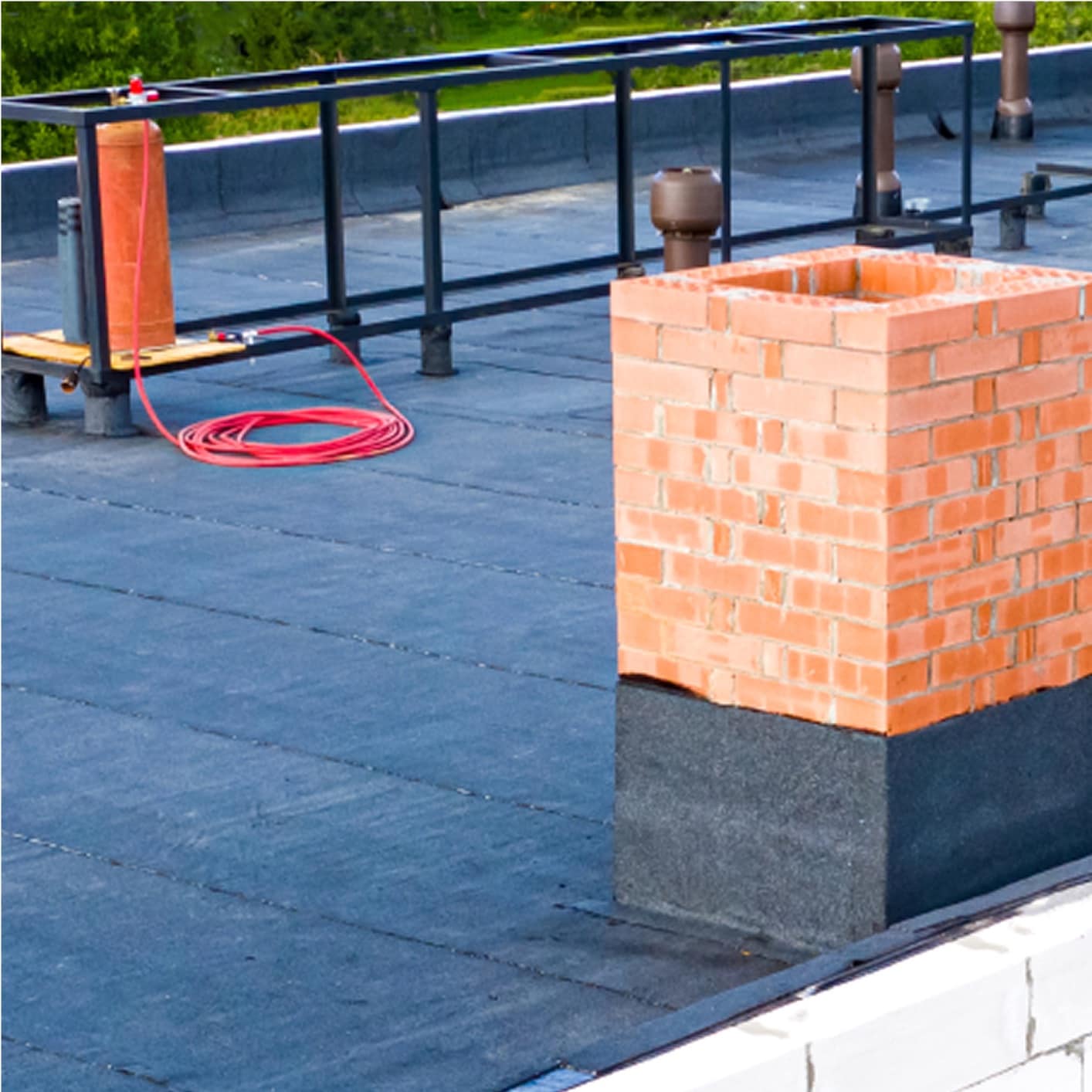Flat commercial roof with exposed black membrane, brick chimney, and ventilation pipes, set against a backdrop of green trees, highlighting roof access and features.
