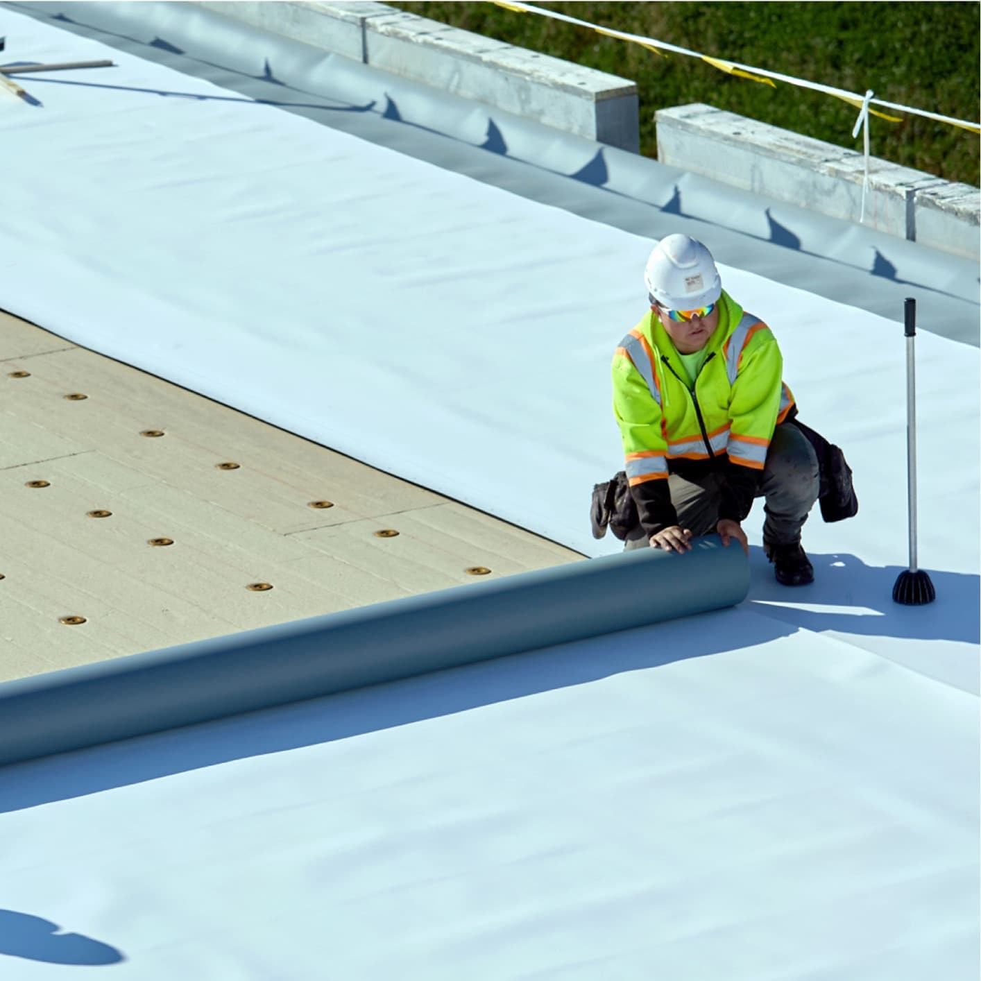 Construction worker in high-visibility gear installing a single-ply roofing membrane on a commercial flat roof, highlighting efficient single-ply roofing application.