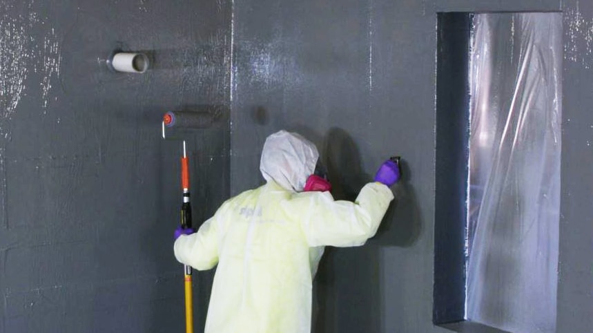 Man in yellow coveralls holding a roller and inspecting a gray wall