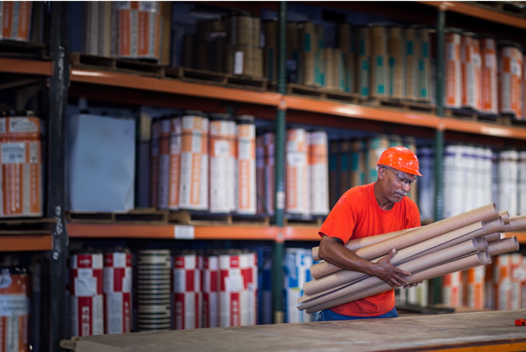 Worker in orange shirt and hard hat carries materials in a Siplast manufacturing facility, showcasing organized product storage and plant tour opportunities.