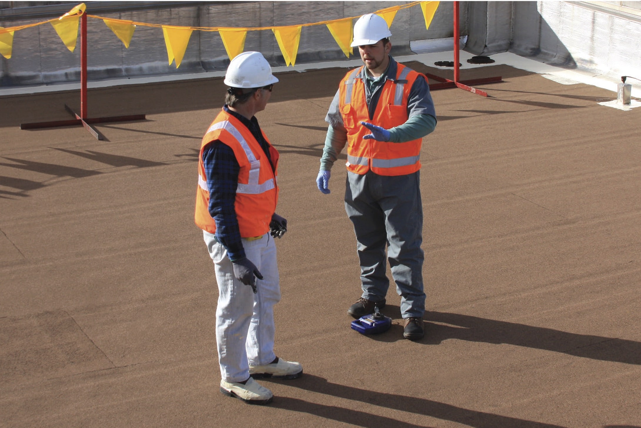Two roofing contractors in orange safety vests and hard hats on a brown Siplast commercial flat roof, discussing the installation or inspection process with safety flagging in the background.