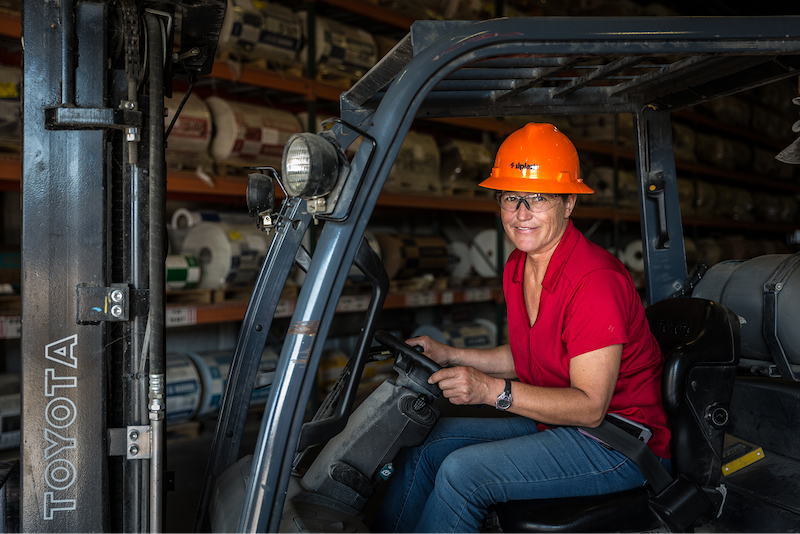 Female worker in an orange hardhat operates a forklift inside a warehouse, symbolizing Siplast's role as industry leaders.