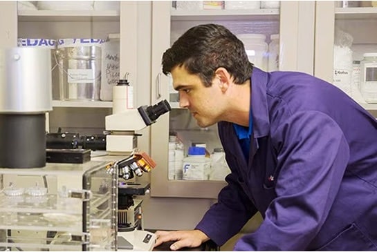A Siplast technician in a lab coat looking into a microscope, representing Siplast's rigorous quality control and the generation of Certificates of Analysis (COA) for roofing materials.