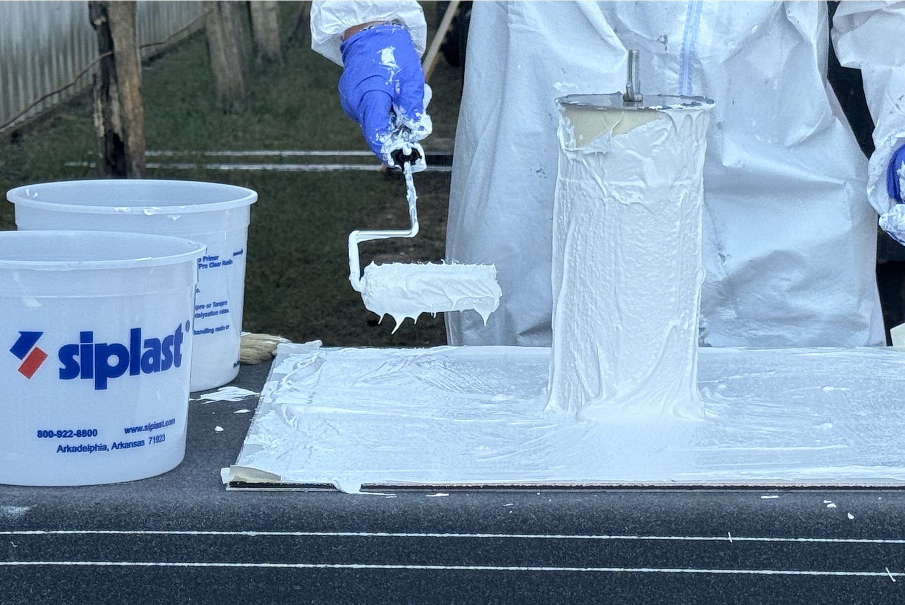 A person wearing protective gear uses a roller to apply a white liquid product to a cylindrical sample, demonstrating a hands-on technical training session for SBS-modified bitumen, PMMA, or other Siplast product applications.