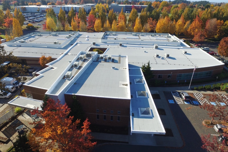 Aerial view of a large modern educational facility with a white roof and surrounding autumnal landscape.