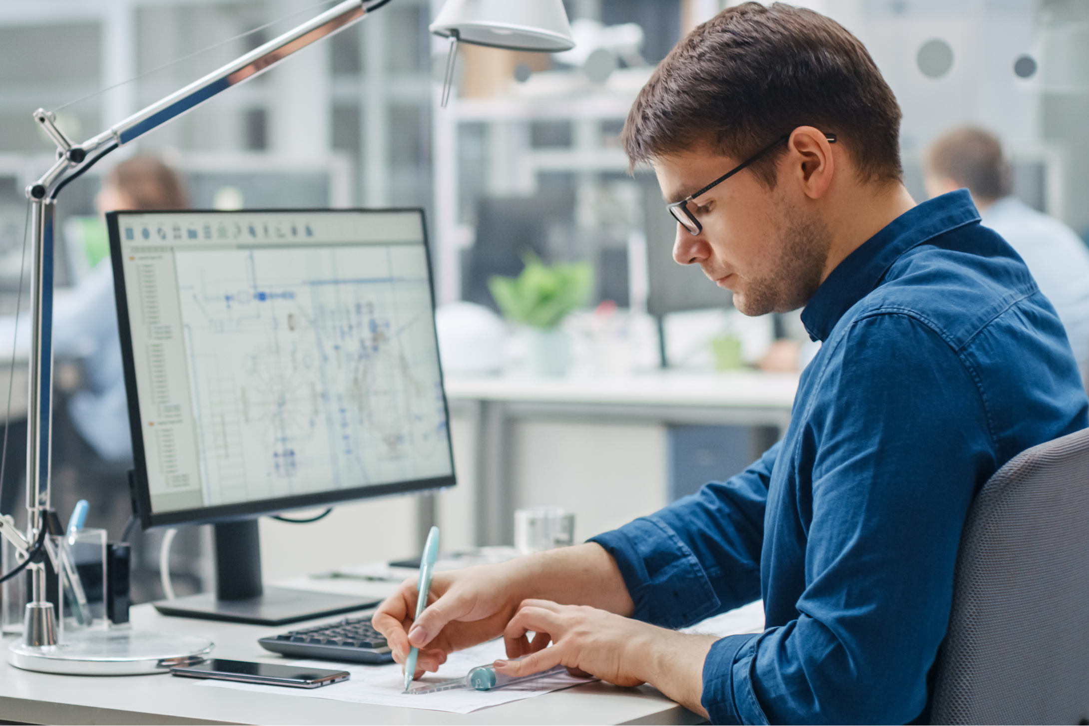 Engineer working at a desk with blueprints and a computer, representing a resource for precise assembly via a detail drawing library.