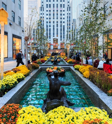 An eye-level view of a bustling urban plaza with a long, narrow fountain featuring bronze sculptures, surrounded by colorful flower displays and pedestrians