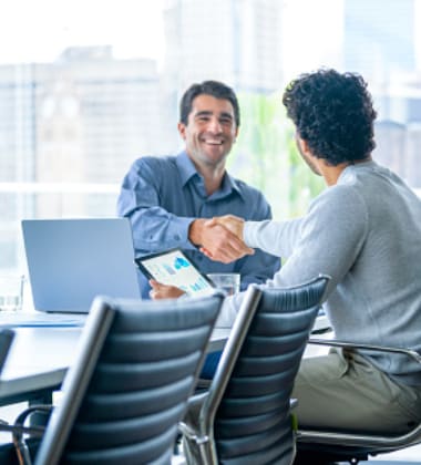 Two men are shaking hands across a table with a laptop and a tablet displaying charts