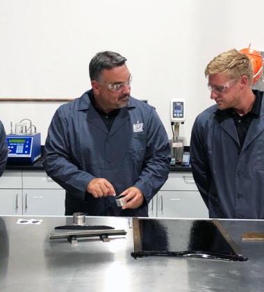 Siplast RISE lab technicians in blue coats and safety glasses conducting material testing on a black roofing sample in a modern laboratory setting, with scientific equipment in the background.