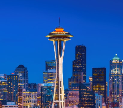 Nighttime panoramic view of the Seattle skyline with the illuminated Space Needle prominently featured against a deep blue sky, surrounded by city lights and other skyscrapers.