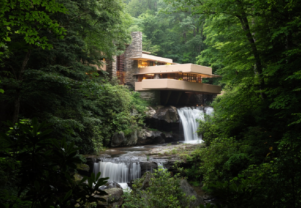Wide view of a modern organic residence situated directly over a rushing tiered waterfall in a lush woodland setting.