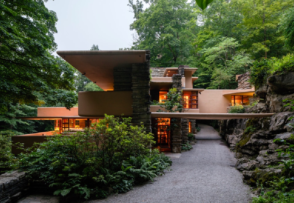 Evening view of a gravel driveway leading toward an illuminated stone and concrete home built into a natural rock ledge.