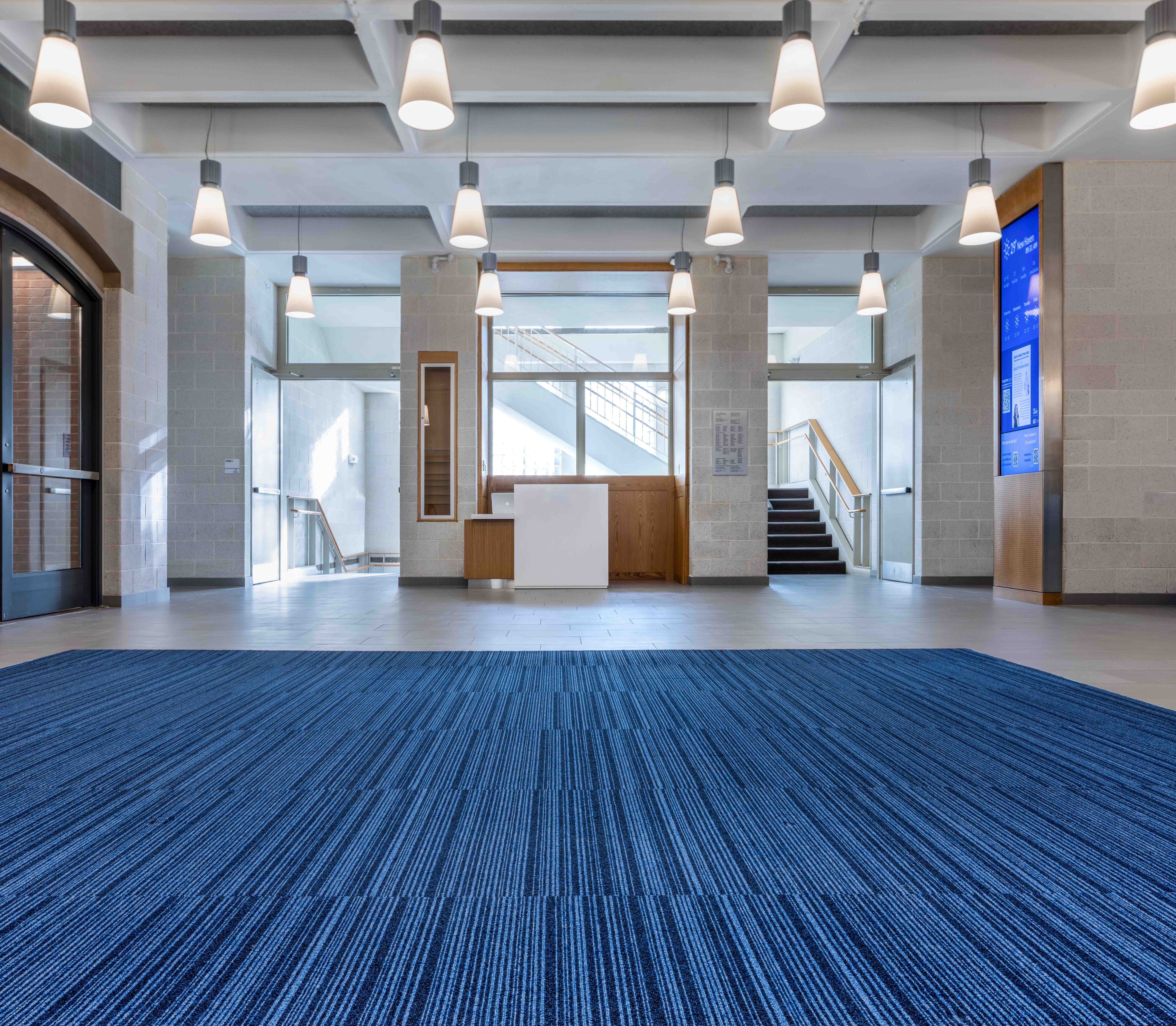 Bright, modern lobby at the Joseph Slifka Center for Jewish Life at Yale featuring blue striped carpet, inviting natural light, and stylish pendant lighting. Staircase visible in the background.