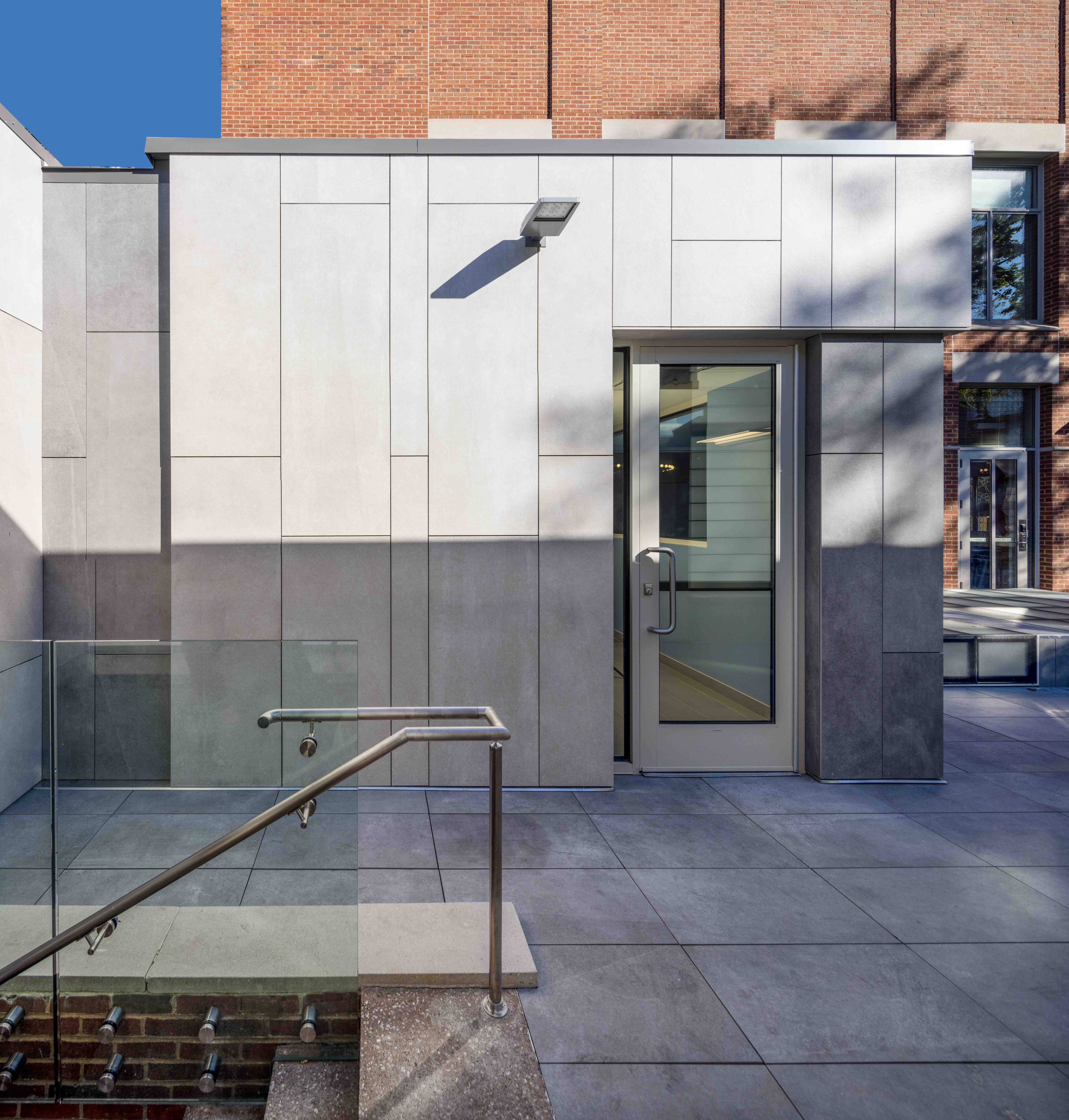 Modern building entrance of the Joseph Slifka Center for Jewish Life at Yale, with sleek stone panels, glass railing, and steps, set against a blue sky and brick wall backdrop.