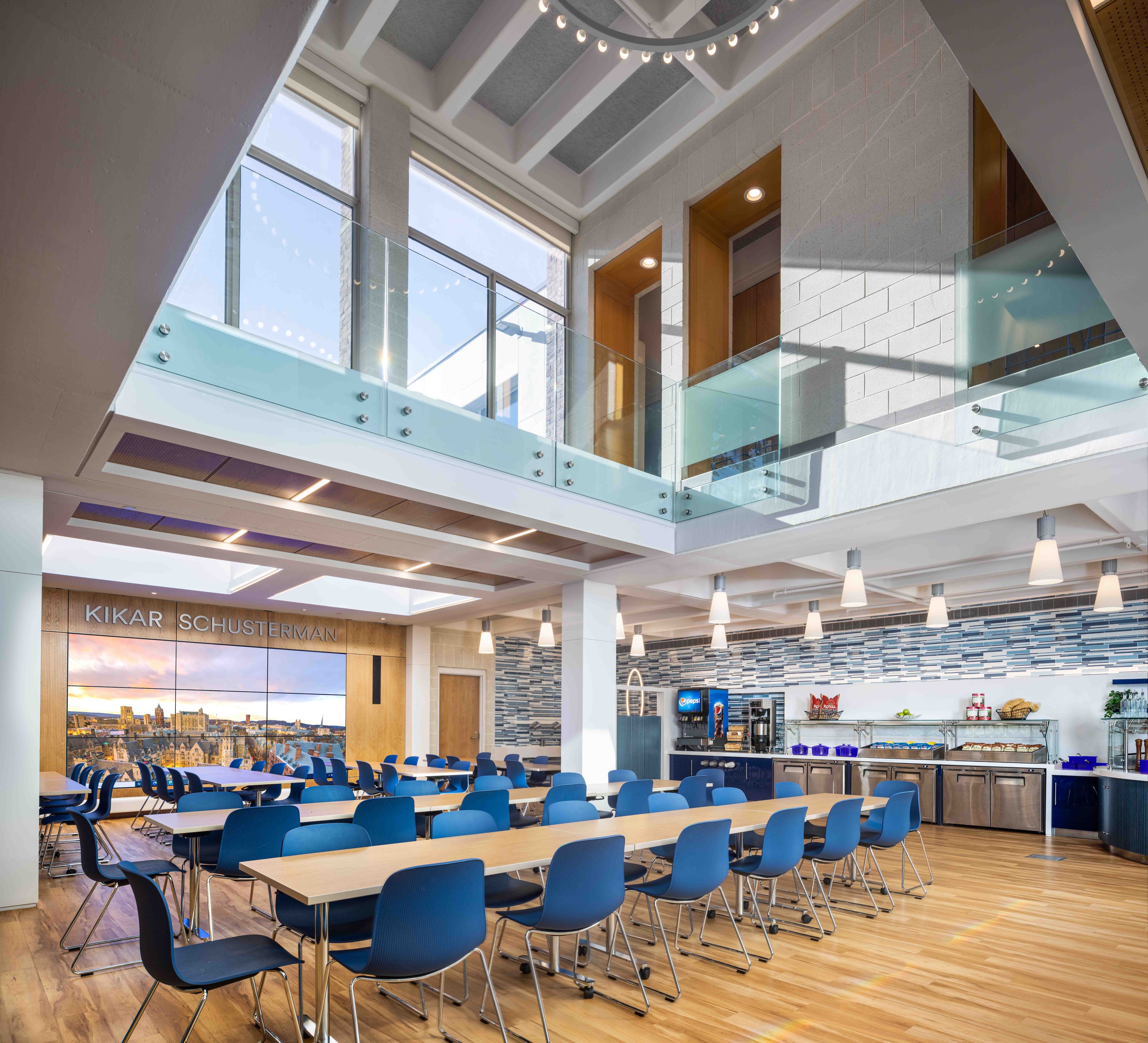 Modern dining hall at the Joseph Slifka Center for Jewish Life at Yale with blue chairs and wooden tables, featuring large windows, a buffet area, and a wall-mounted screen displaying a city view.