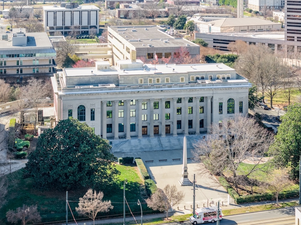 Wide aerial shot of a majestic government edifice fronted by a stone obelisk monument and a wide concrete plaza.