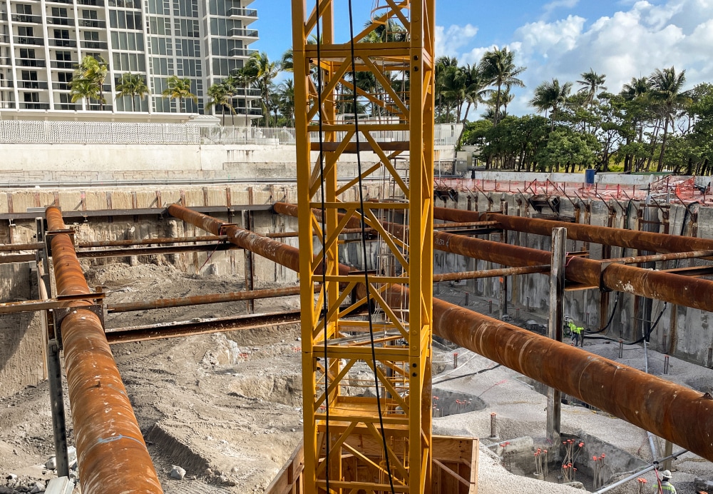 Aerial view of a deep foundation excavation site featuring massive horizontal steel bracing and a central yellow crane tower near the Florida coastline.