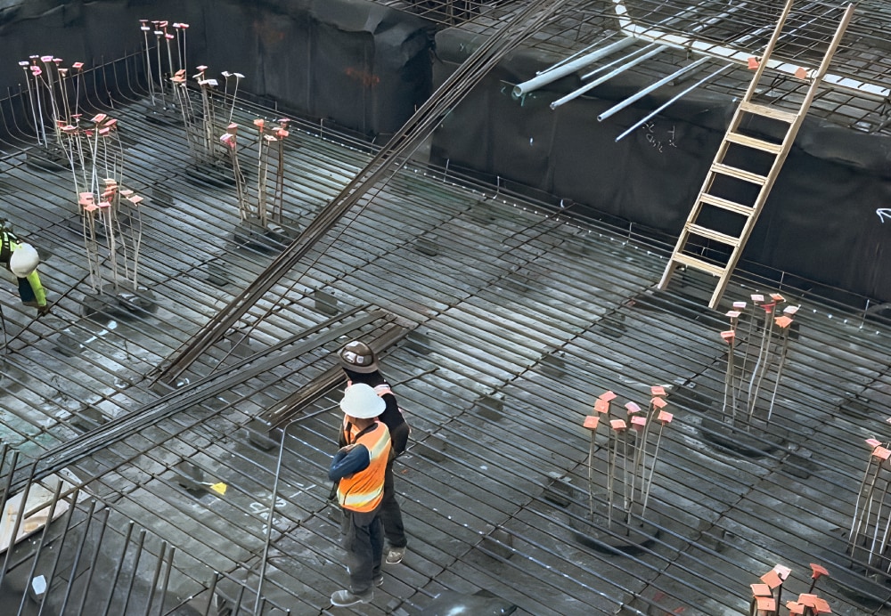 Overview of a dense steel rebar reinforcement grid being laid over a waterproofed foundation floor, showing workers coordinating the structural assembly.