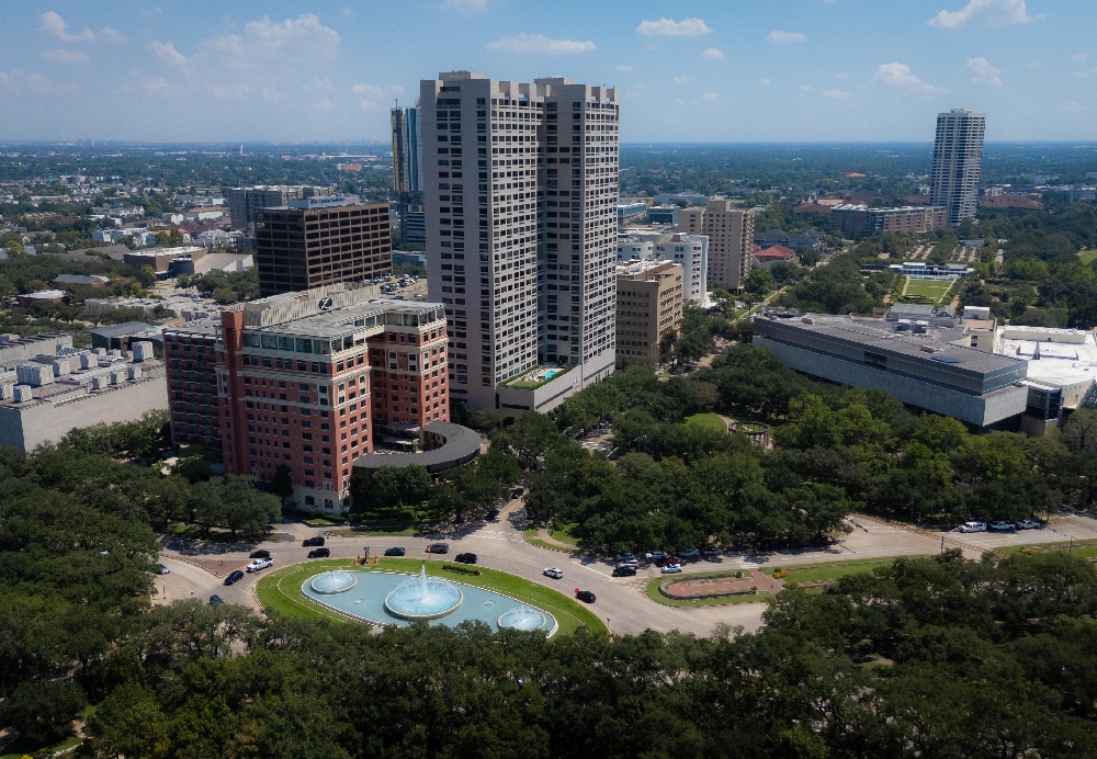 High-angle cityscape showing a brutalist-style residential tower nestled among lush green trees near a public park fountain.