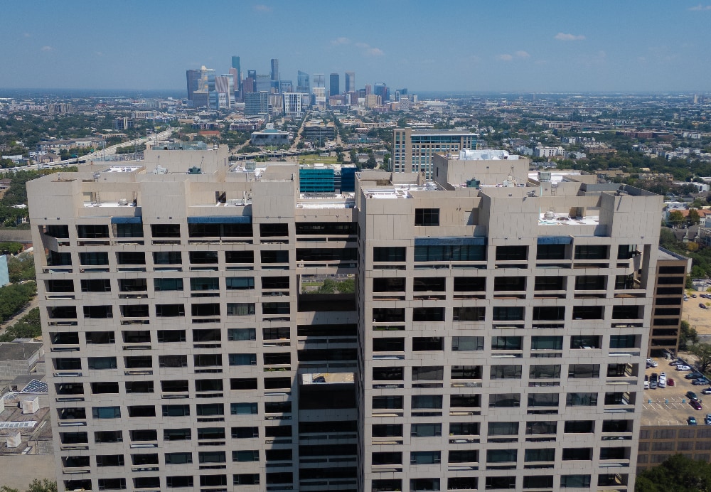 Front-facing view of a concrete residential high-rise with the distant Houston downtown skyline visible in the background.