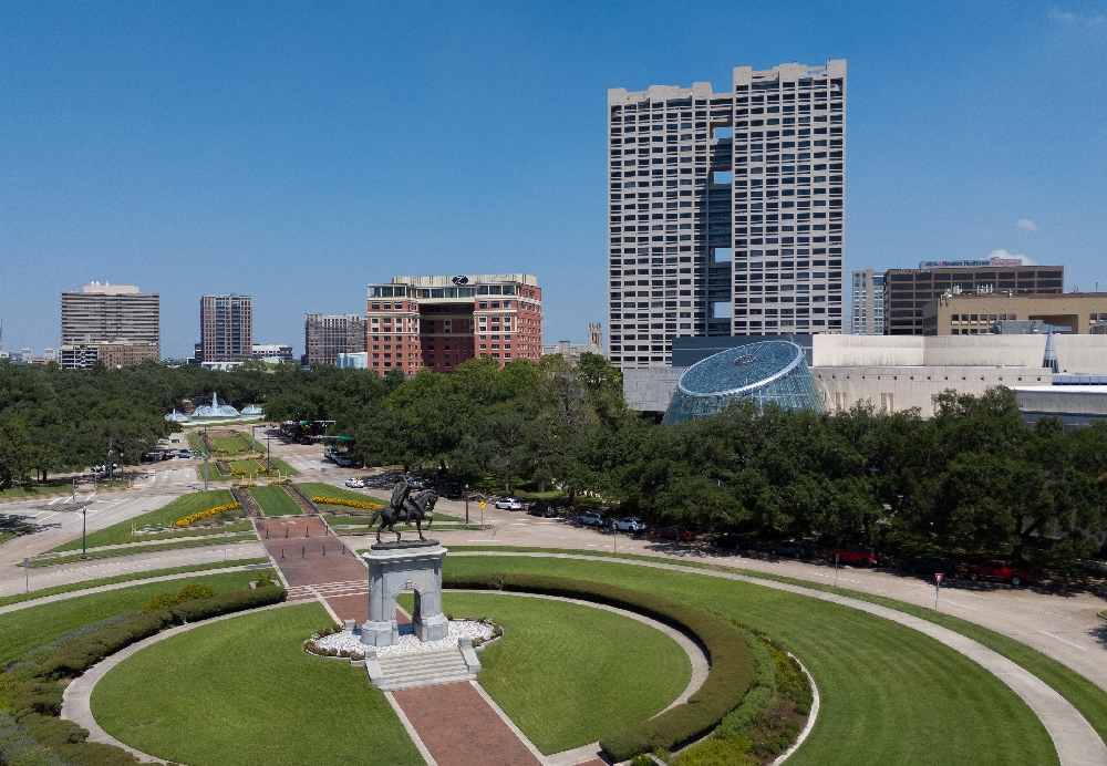 Ground-level panoramic view of an urban roundabout with a bronze statue and a towering residential landmark under a clear blue sky.