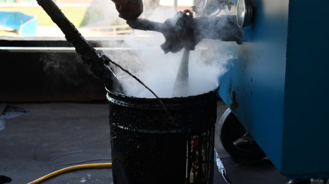 A hand holding a tool stirring hot, steaming black asphalt material in a black bucket next to a blue melting machine.