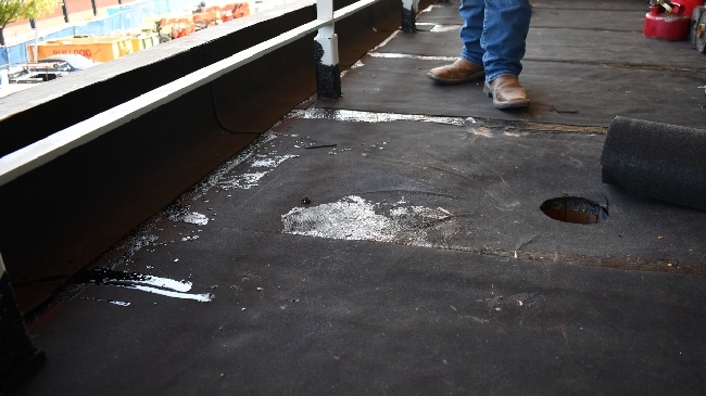 A close-up of a dark roofing membrane with visible adhesive and a circular opening, with a person's feet in work boots in the foreground.