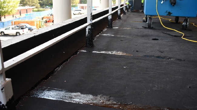 A close-up of a dark roofing membrane showing applied adhesive along edges and around a white railing post, with liquid pooling in some areas.