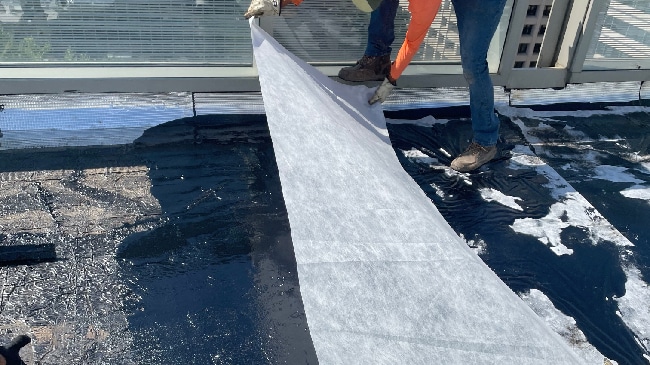 A construction worker unrolling a white protective fleece onto a dark, rubberized asphalt roofing layer.