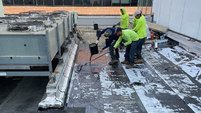 Multiple construction workers in safety vests applying hot liquid asphalt from buckets onto a partially completed rubberized roofing system near HVAC units.
