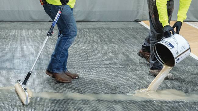 One man pours Pro Primer Resin onto a concrete surface while another man uses a roller to spread it