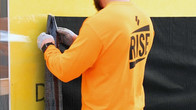 Worker installing a black Siplast self-adhered air barrier membrane onto yellow wall sheathing.