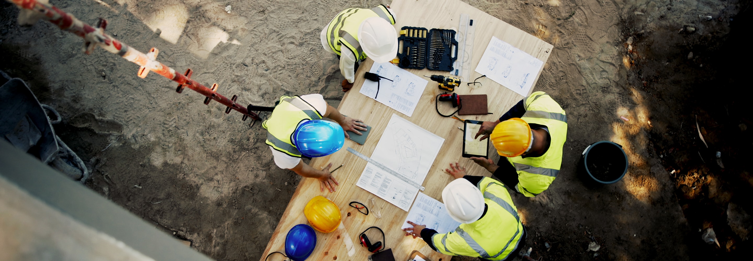 Overhead view of construction workers reviewing blueprints and a tablet on a job site, highlighting educational resources.