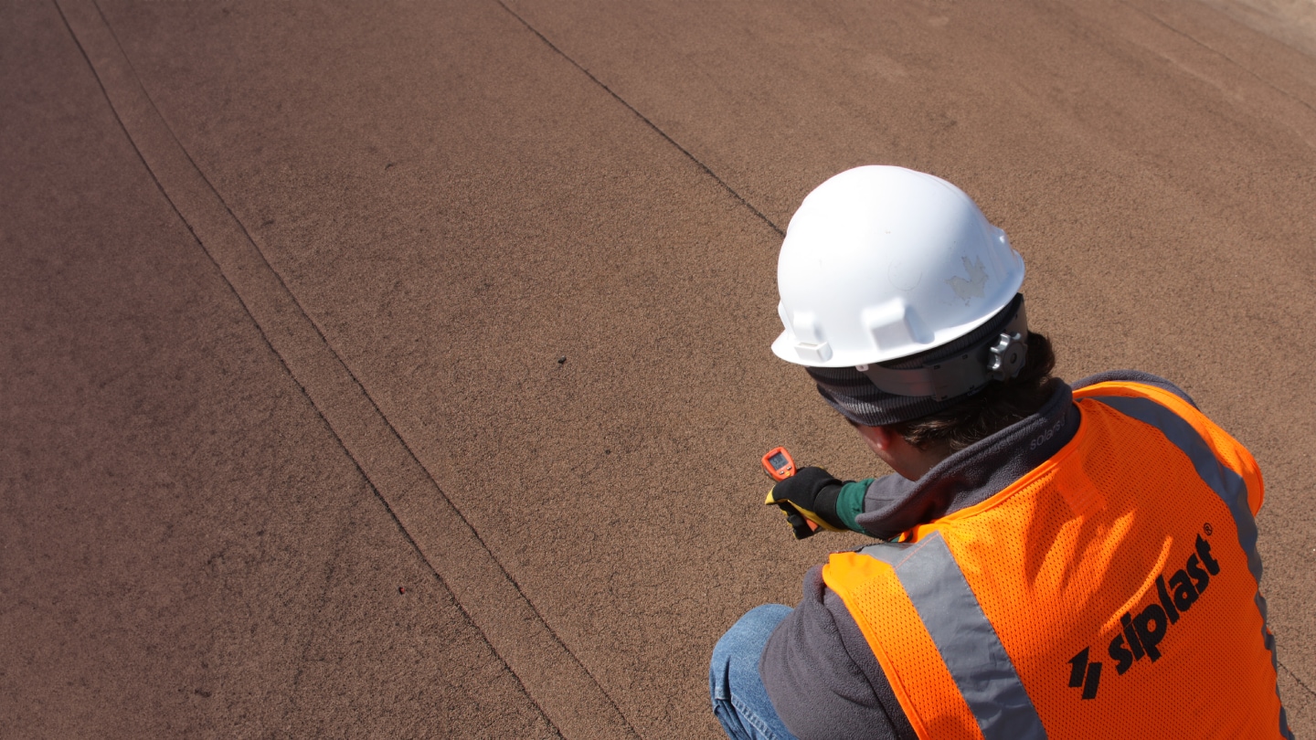 A roofing professional in a hard hat and safety vest inspecting a Siplast roof.