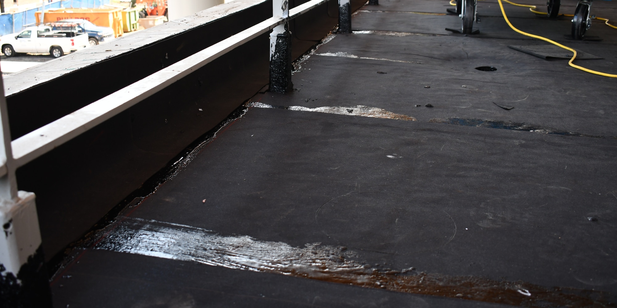 A close-up of a dark roofing membrane showing applied adhesive along edges and around a white railing post, with liquid pooling in some areas.
