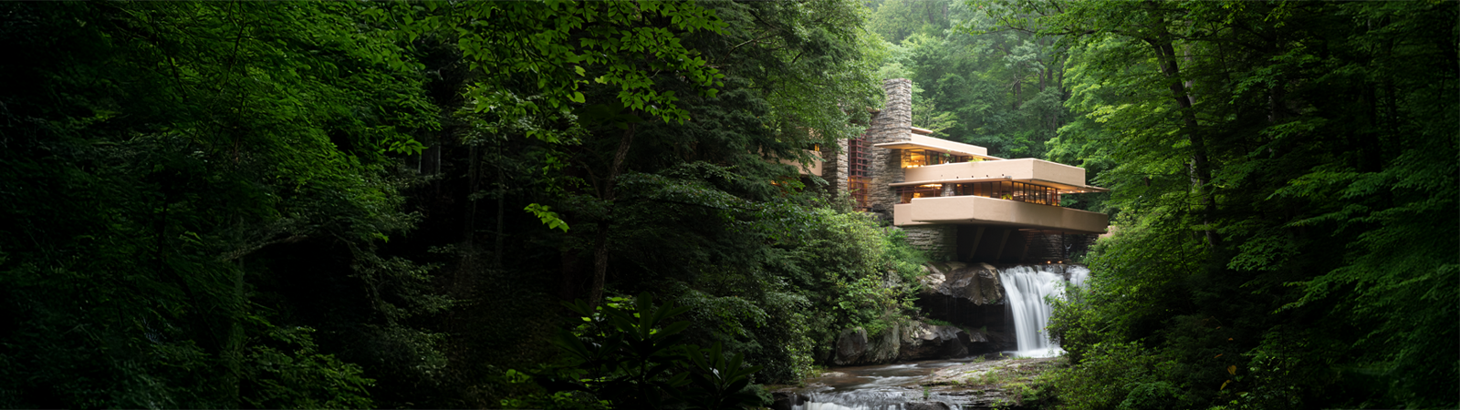 Panoramic view of the organic modern architecture with beige cantilevered terraces extending over a natural waterfall surrounded by lush green summer forest.