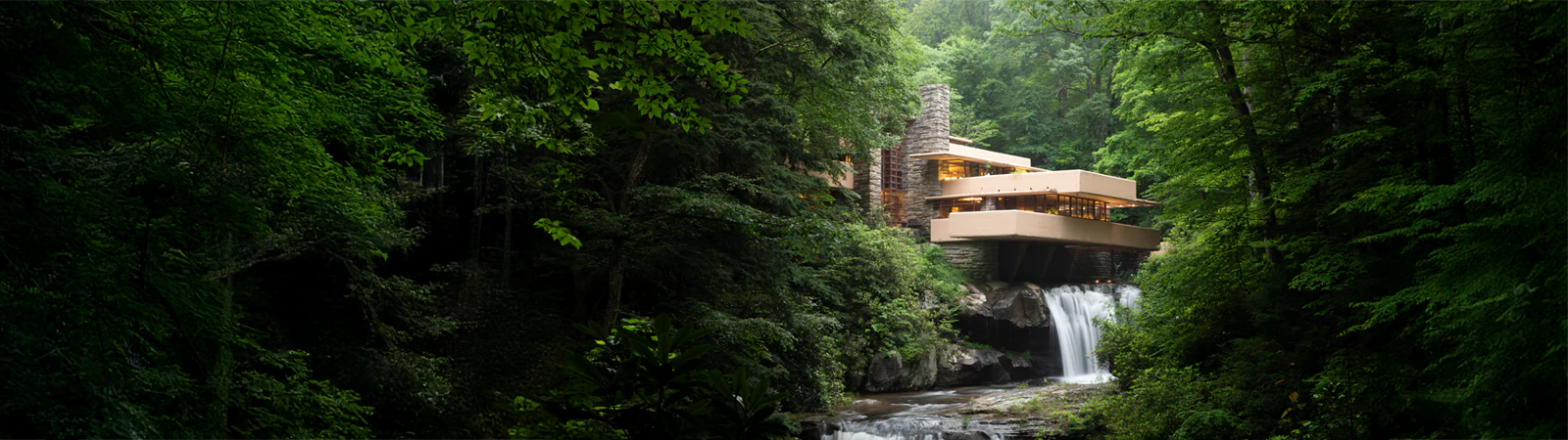Panoramic view of the organic modern architecture with beige cantilevered terraces extending over a natural waterfall surrounded by lush green summer forest.