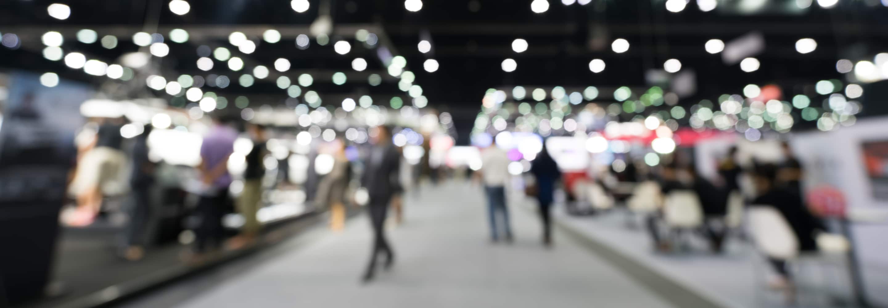 Blurred background of a busy, well-lit trade show floor, highlighting the section for finding Upcoming Industry Events.
